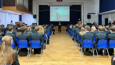 Olivia Richwald/BBC Rows of bright blue plastic chairs  separated by a central aisle. The chairs are mostly occupied by students wearing dark blazers, all facing forward toward a stage area. At the front, a male student is stood holding a sheet of green paper. Behind him there is a large projection screen with the image of a man and words on.