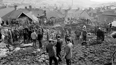 Mirrorpix via Getty Images The Aberfan disaster - crowds of men look on in this black and white photograph standing on the rubble with a digger visible in the background