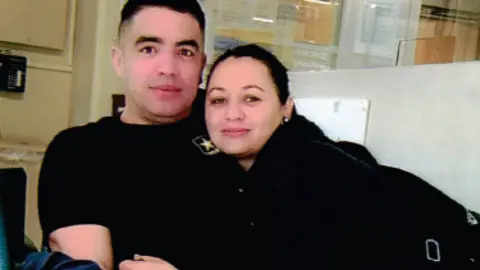 Sgt First Class Jose Serrano and his wife, Deisy Rivera Ortega, who is leaning on his shoulder. Both wear black. Sgt Serrano wears a t-shirt with the US Army logo.