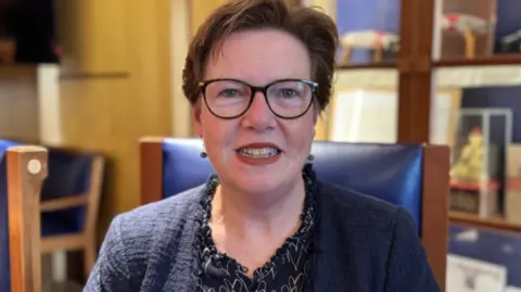 Jane Poole-Wilson, a woman with short brown hair and black glasses, she has lipstick on, and is wearing a navy cardigan over a black and white blouse.