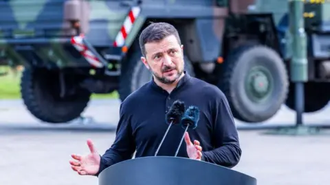 Volodymyr Zelensky, President of Ukraine, speaks into a microphone at a podium wearing a black shirt. Behind him is a military vehicle.