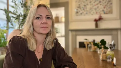 Chloe has shoulder-length blonde hair and is seated at a wooden table in a softly lit café, wearing a brown long-sleeved dress. Plants, glassware, and framed artwork decorate the warm, modern interior.