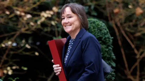 TOLGA AKMEN/EPA/Shutterstock Jo Stevens is photographed arriving at 10 Downing Street for a cabinet meeting in London. She is holding the red budget box in her hand. She is wearing a navy-blue blazer and a top with a floral pattern.