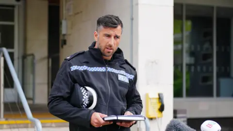PA Media Superintendent Matt Ebbs wears a police uniform, holding his hat under his arm. He holds a written statement as he reads to the press standing outside a building.