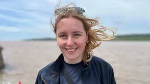 A young woman with blonde hair looks at the camera. She wears a navy top and hoody and has sunglasses on her head. She is standing in front of a body of water. 