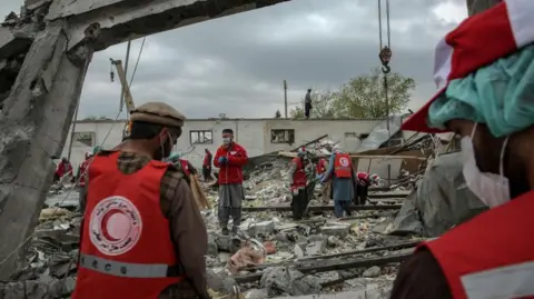 EPA/Shutterstock Several volunteers from the Red Crescent remove debris from the destroyed rehab centre in Kabul. There are about 13 men (some partially obscured by others), wearing red tabards with the organisation's crescent logo and medical face masks.