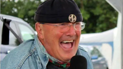 BBC Dewi Morris laughing into a microphone, wearing a beanie hat, and glasses