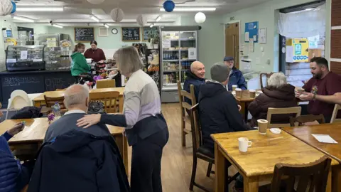A cafe with lots of customers drinking hot drinks. A woman with grey hair stands with her arm around a man sitting at a table who is taken from behind. Another table has five people sitting around it. They are chatting and smiling.