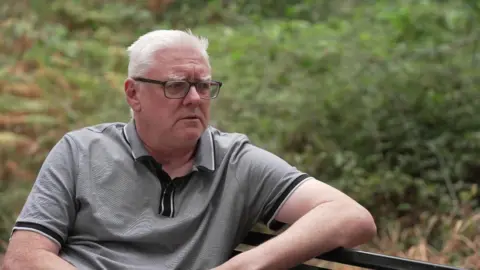 A man with white hair and glasses sat on a bench and staring off the camera