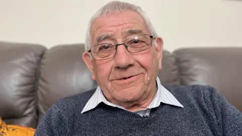 Hugh Fraser sits in a large brown leather armchair and looks at the camera. He is wearing a blue jumper and open necked shirt. He wears glasses with thin black rims. He has thinning grey hair, flattened down at the front.