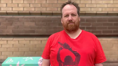 Adam is wearing a red t-shirt and is standing in front of a wall and a painted junction box which has been painted light green and the covered in light pink symbols like stars and hearts. Adam has light brown receding hair and ginger coloured beard.