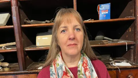 John Devine/BBC Jan Summerfield has light brown, shoulder length hair and is wearing a maroon woollen jumper with a silk scarf with floral designs on it. Behind her is a dark wood shelf unit with several small compartments.