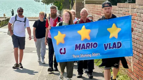 A group of around a dozen walkers are heading up a slope holding a blue banner with three yellow stars and the names CJ, Mario and Vlad. The sun is shining and there is a shingle beach and the sea in the background.
