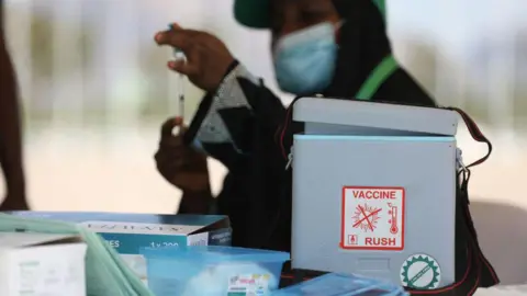 Getty Images This image shows a vaccine storage and preparation area, with a portable insulated cooler prominently positioned in the foreground. A label on the cooler reads “VACCINE” and includes a temperature graphic, indicating the importance of maintaining a controlled cold chain. Nearby are medical supplies, including boxes, trays, and packaged equipment arranged on a work surface. In the background, a person wearing a face mask is holding a syringe upright, appearing to prepare or inspect it before use. The shallow depth of field keeps the cooler and vaccine-related equipment in sharper focus while the background remains blurred. The overall scene emphasizes the handling, storage, and preparation of vaccines in a healthcare or immunization setting.