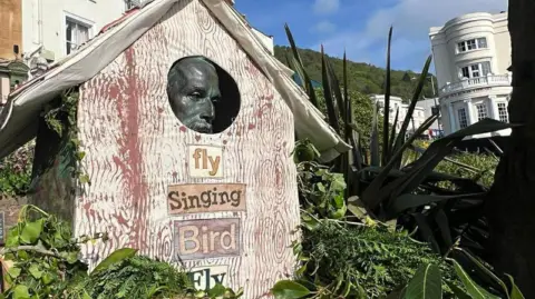 Kirsty Johnson A wooden bird house surrounds a stone head. on the front of the box it says "fly, singing bird, fly". there are green plats around it