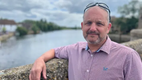 Head and shoulders image of Steve Hodgson. Steve is wearing glasses on the top of his head and has a grey closely shaved beard. He is wearing a short-sleeved shirt and is leaning his arm against a wall with a river behind him.