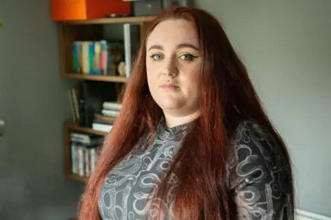 A woman with long dyed red hair and blue eyes stand in her living room. There is a bookshelf behind her. She has a grey top on. It is a head and shoulders shot.