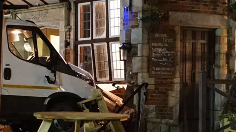 A white vehicle crashes into a restaurant. The windows are buckled and wooden furniture outside the building is damaged.
