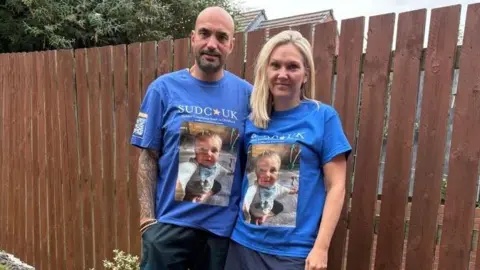 FAMILY HANDOUT A man and a woman stand next to a garden fence, wearing blue charity fundraising t-shirts showing the face of their late son
