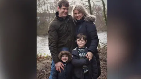 Rosie Moss Rosie and Ben stand with two of their children who are young boys. They're all wearing warm coats and are standing in front of a river.