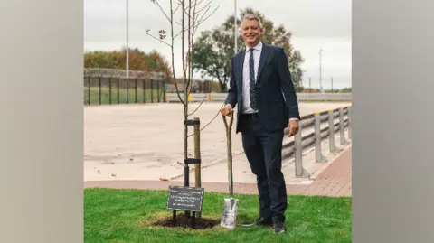Luke Pollard in a suit, standing with a shovel on a patch of grass, next to a sapling and a small, black plaque on the ground.