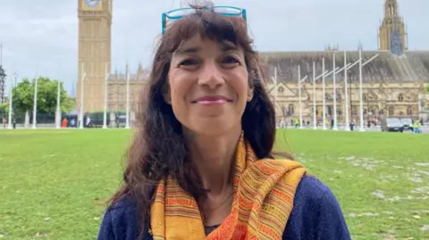 Lib Dem MP Manuela Perteghella standing outside the Palace of Westminister. She has long brown hair, has blue-rimmed glasses on her head and is wearing a yellow scarf and a blue top.