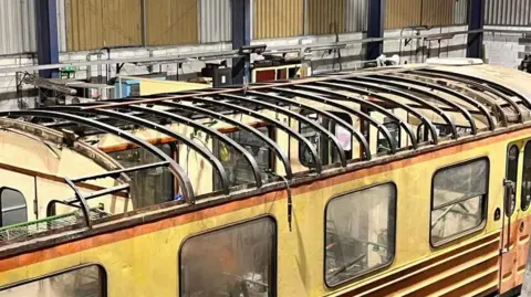 Nene Valley Railway The carriages from the above image are inside what looks to be a warehouse. They are missing roofs - instead only the rounded metal framework for a roof can be seen. The orange and yellow body of the railcar is dirty.
