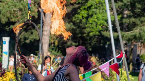 Steamship Circus A woman with pink hair is picture in profile looking up into the sky and breathing a burst of fire into the air. In her hand she is holding a lit torch. The picture is taken outside on a sunny day and spectators can be seen in the background. 