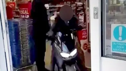 A man wearing black rides on a moped inside a convenience store. He is seen through the entrance doors as he rides towards them. A stack of baskets and a security guard can be seen.