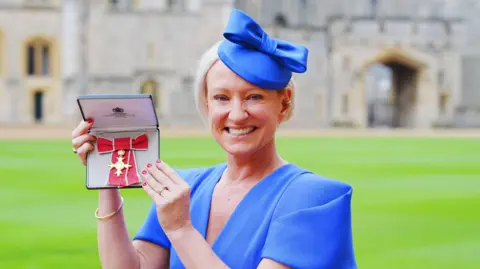 PA Media Ruth Dodsworth stands wearing a blue blouse in palace grounds smiling at the camera.