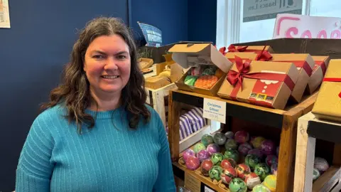 Martin Heath/BBC Sarah Prudames with long dark hair smiling at the camera while wearing a blue cardigan with shelves of colourful soaps behind her.