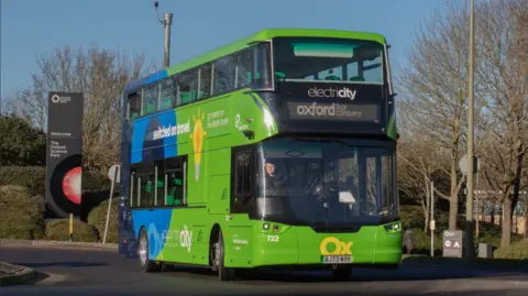 A green electric bus labelled Oxford Bus Company drives out of Oxford Science Park.