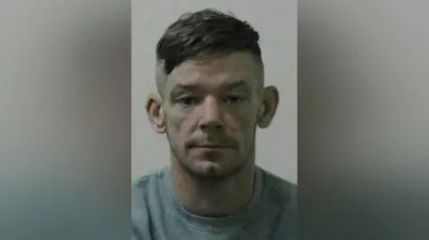 Northumbria Police Mugshot of Ness. He has short dark hair with shaved sides and a stubbly beard. He is wearing a grey sweatshirt