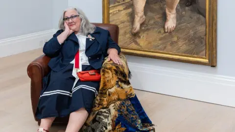 Sue Tilley is wearing a navy knee-length skirt, a navy jacket, and white blouse. She has a small bright red crossbody bag and a decorative brooch pinned to her lapel, and red lace-up shoes with white soles. She is resting one arm on the chair and supporting her head with her hand and smiling. She has glasses and has shoulder-length grey hair.