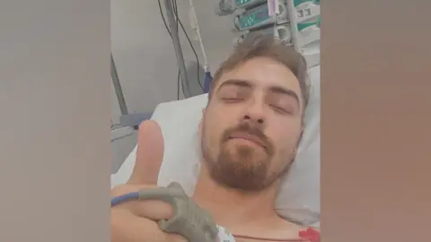 Liam Watson A man with short brown hair and a beard is lying in a hospital bed giving a thumbs up in a selfie. He has wire attached to his chest and is wearing a monitor on his finger.