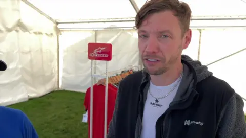 Toby Lambert from Alton Sports giving an interview after news of the event's cancellation. There is a rack of clothes behind him. He is under a tent. Another person could be seen near him.