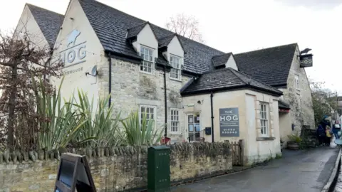 A white public house building with a sign on the side of the building stating its name "The Hog" - there is a pavement in front of the building and some bushes in the foreground.