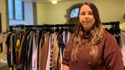 Rosemary smiles at the camera with the clothes rail behind her. She has a brown turtle neck jumper on and has long brown hair.