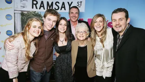 Getty Images Seven people stand with their arms around each other's shoulders in front of a partition with the graphic of a street sign saying "Ramsay St"