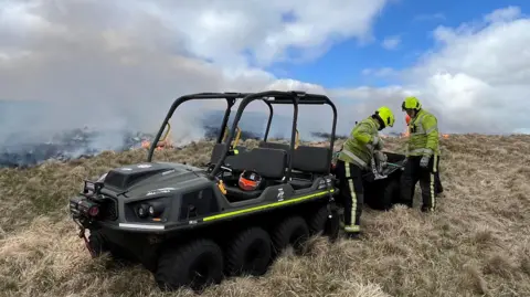 A mobile 'buggy' and two firefighters next to it focussing on something. There is smoke behind them 