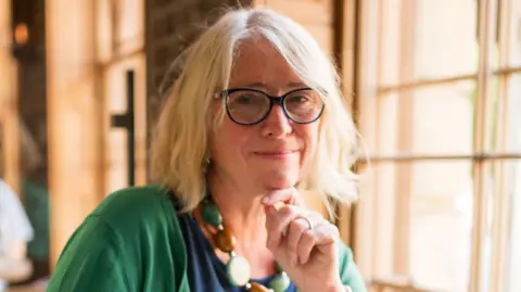 Deborah Humphrey A woman with blonde hair and glasses looks at the camera and touches her chin. She is wearing a navy top, green cardigan, and a necklace of large green and brown circles, and is sat indoors near a window.