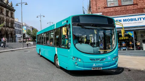 Sarah Caldecott A blue Arriva bus waits on a wide corner outside a mobility shop. There is no destination banner up but a driver is sat inside the bus. 