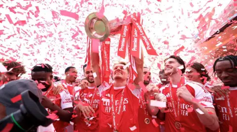  An Arsenal player lifts the Premier League trophy above his head as red and white confetti falls around him, with teammates in red shirts celebrating alongside.