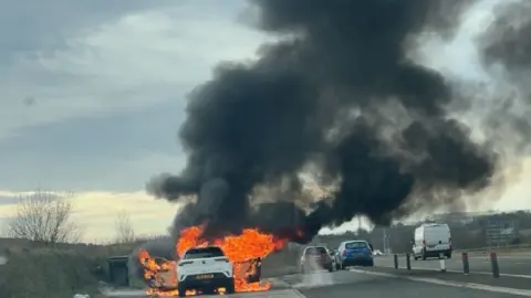 Alex McClean A white car engulfed in orange flames with a large black cloud of smoke rising in the air. The car is parked in a lay-by while other cars are still driving on the road.