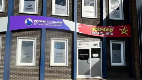 Gary Johannes Exterior of the building- with a purple and white Inspired to change board and a white door and white windows.
