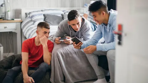 Three teenage boys in a bedroom sitting on and around a bed. One of them is holding a PlayStation controller, while another shows his friends something on his smartphone.