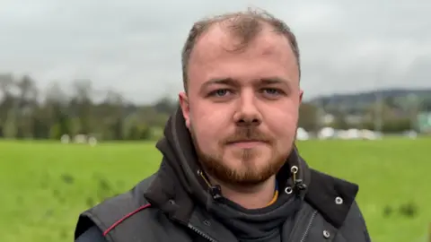 A man with some hair and a reddish beard standing outside in front of a field. He is wearing a black jacket.