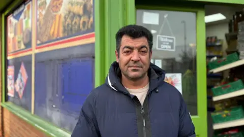 Mr Ali standing outside the Sultan Halal Superstore. The shop is painted green and has pictures of food in the windows. Mr Ali has black hair and stubble and is wearing a blue coat.