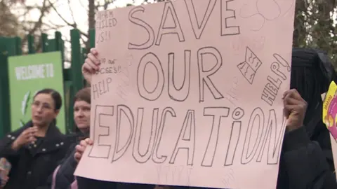 A banner on pink paper or cardboard that says " Save our education" that is being held up by pupils, who can be seen behind it.