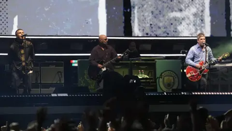 Jordan Pettitt/PA Wire Oasis on stage at the Principality Stadium, Cardiff, as the band's long-awaited reunion tour kicks off in Wales. A man leans into the microphone while singing and another man plays the guitar with another man on the right playing a red guitar and wearing a denim shirt. The hands of the crowds can be seen raised in front of the stage.